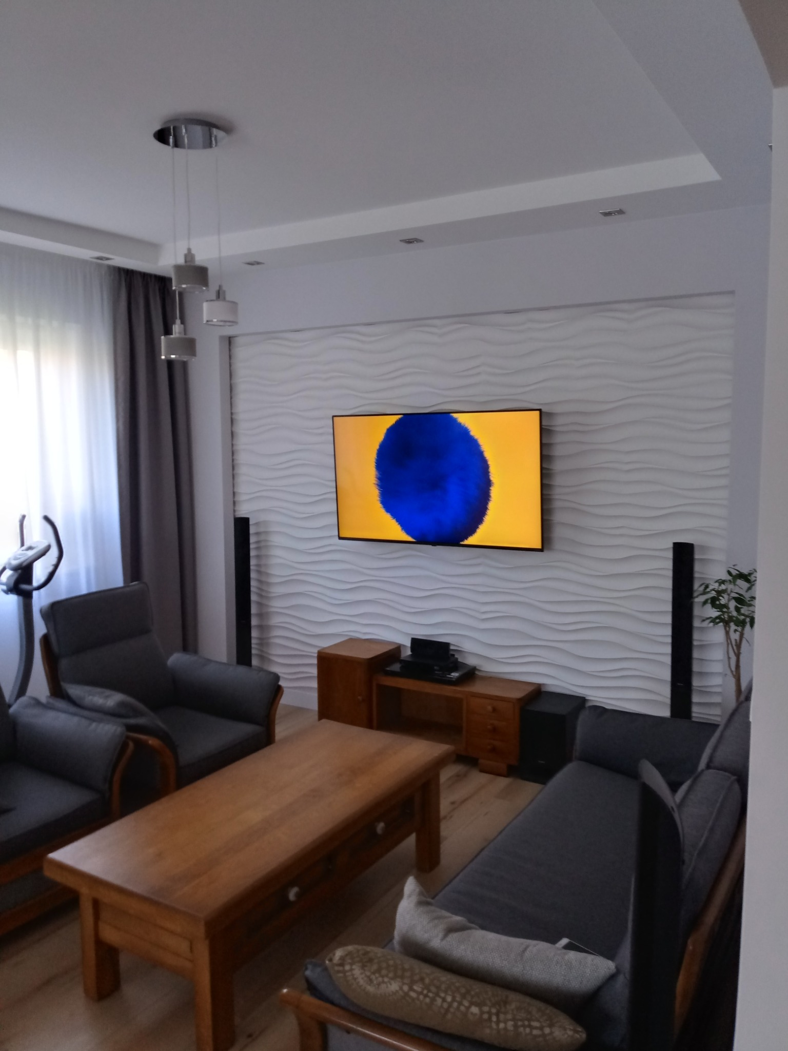 Nowoczesny salon z teksturowaną ścianą 3D, telewizorem, szarą sofą i drewnianym stolikiem kawowym oraz lampami wiszącymi.