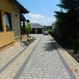 Szeroki podjazd z kostki brukowej w dwóch odcieniach szarości i beżu prowadzący do garażu, obok domu z oknami w czarnych ramach i zadbanego żywopłotu z małymi drzewkami iglastymi.