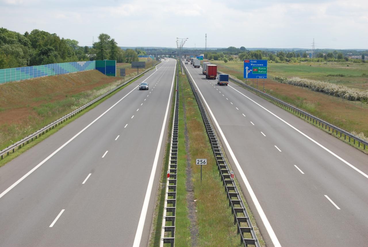 Autostrada A2 z widocznymi ciężarówkami w oddali, znakami drogowymi wskazującymi kierunek na Łódź i Warszawę, oraz samochodem osobowym jadącym lewym pasem.