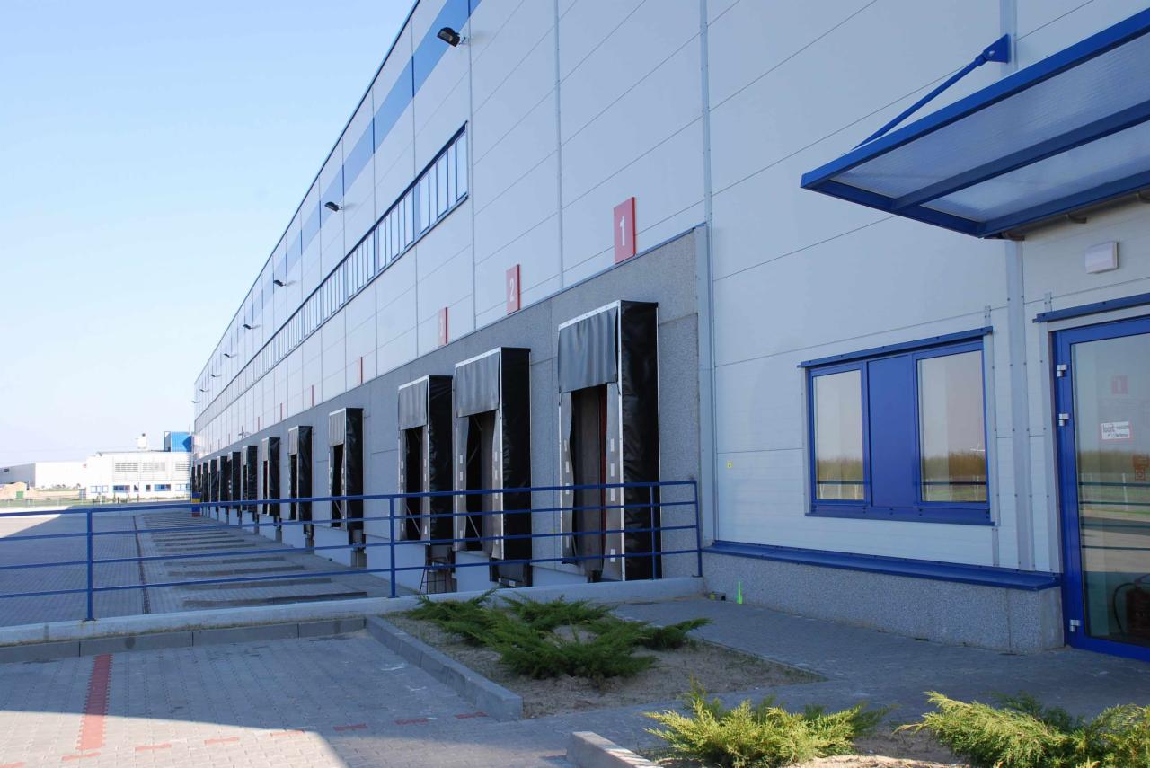 Rampa rozładunkowa magazynu logistycznego z niebieskimi akcentami, widok z zewnątrz w słoneczny dzień.