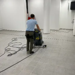 Mężczyzna czyści szarą, kafelkową podłogę w jasnym pomieszczeniu za pomocą przemysłowej maszyny czyszczącej Karcher, widoczny kabel zasilający i biały filar w tle.