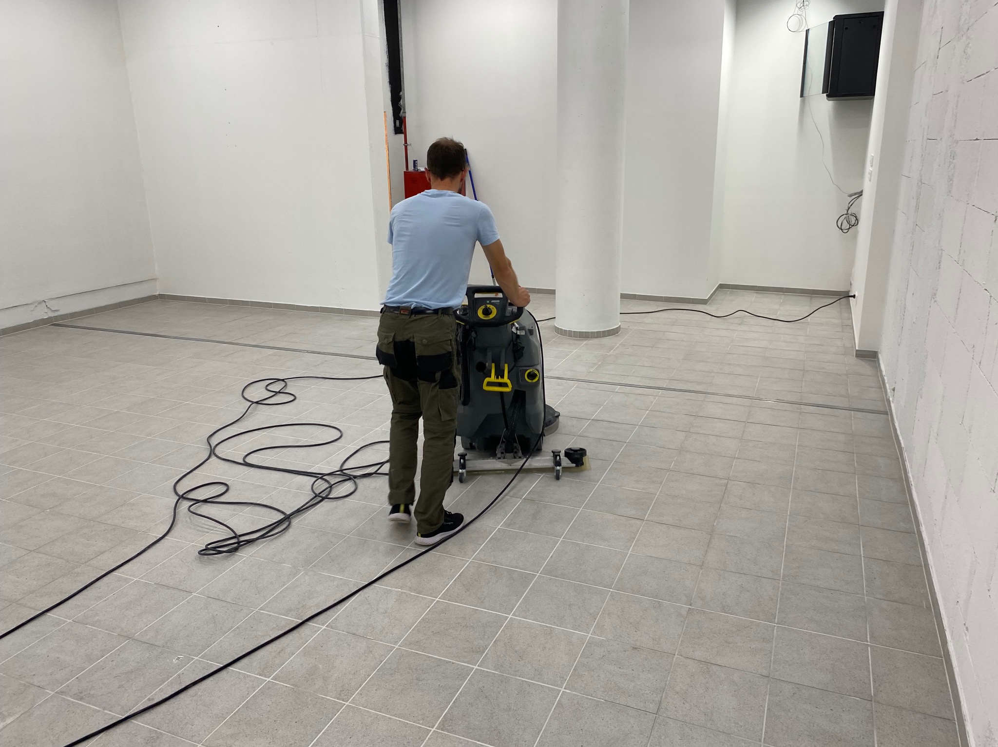 Mężczyzna czyści szarą, kafelkową podłogę w jasnym pomieszczeniu za pomocą przemysłowej maszyny czyszczącej Karcher, widoczny kabel zasilający i biały filar w tle.