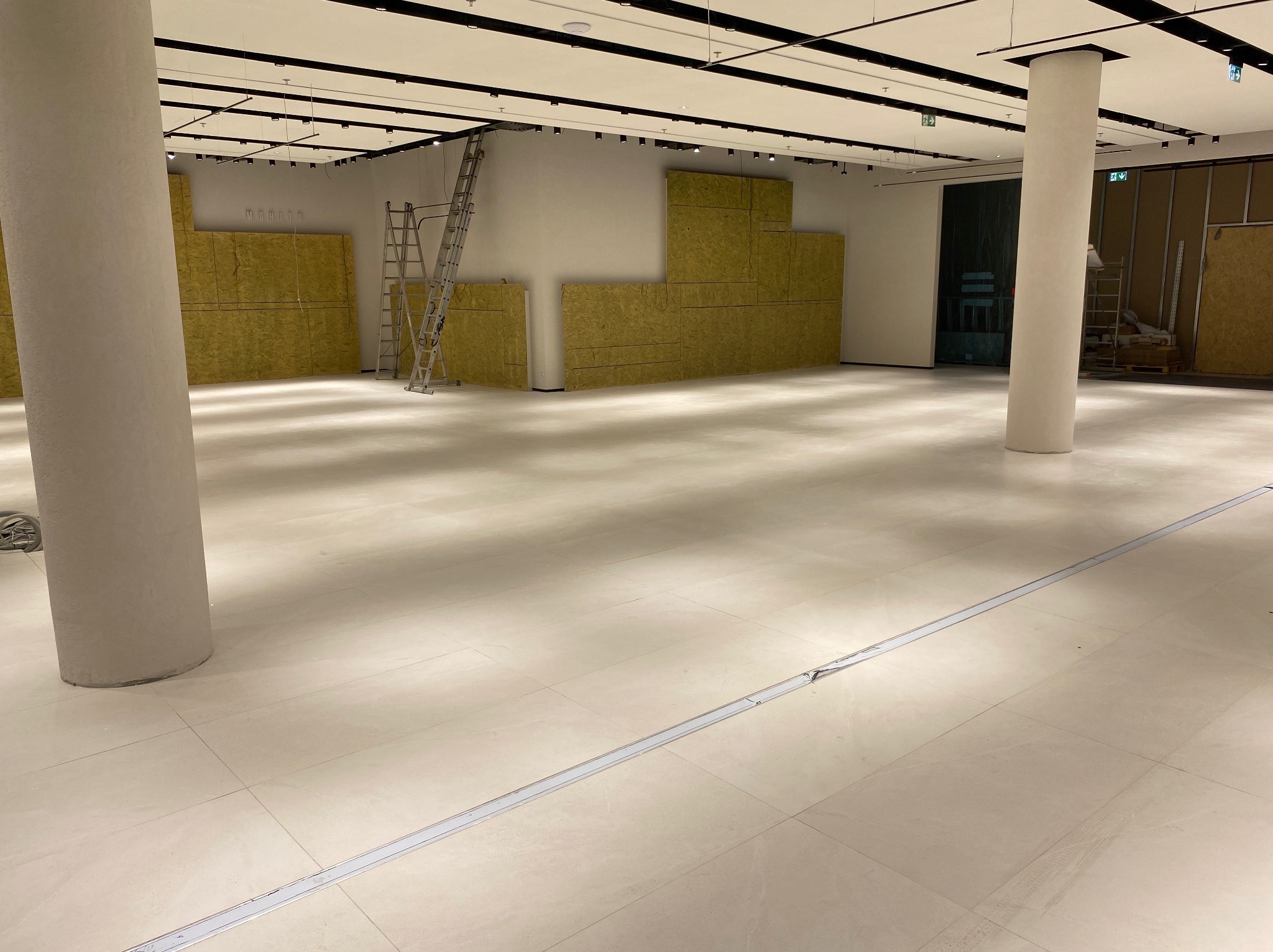 Wnętrze sklepu w trakcie wykańczania: położona jasna podłoga z płytek, obudowane filary, ściany wyłożone panelami izolacyjnymi, drabiny i narzędzia w tle.