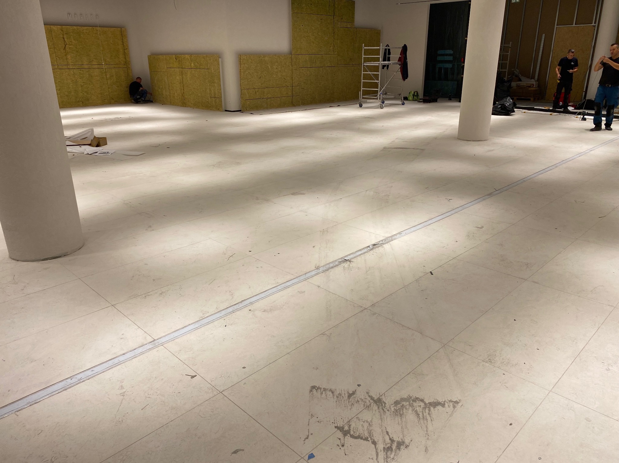 Wnętrze w trakcie prac wykończeniowych: położona podłoga z płytek, zabezpieczona szczelina dylatacyjna, ocieplenie ścian wełną mineralną, pracownicy w tle.