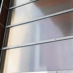 Zadaszenie balkonu wykonane z przezroczystego poliwęglanu komorowego, mocowane do pomarańczowej ściany budynku za pomocą metalowych profili.