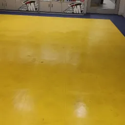 Żółta podłoga z płytek w sklepie, z widocznymi śladami użytkowania i zabrudzeniami, przylegająca do granatowej podłogi w tle, widoczny fragment regału sklepowego i kosz na śmieci.