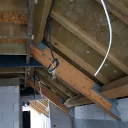 Surowy stan poddasza w Olsztynie z widoczną izolacją, drewnianą konstrukcją dachu i prowizorycznym oświetleniem w postaci żarówki wiszącej na kablu, oraz częściowo poprowadzonymi przewodami...