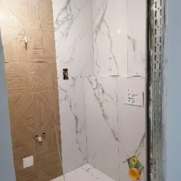 Wnętrze łazienki w trakcie wykańczania: ściany wyłożone białymi płytkami imitującymi marmur, fragment ściany z drewnopodobnymi płytkami z geometrycznym wzorem, widoczne puszki elektryczne i rury...