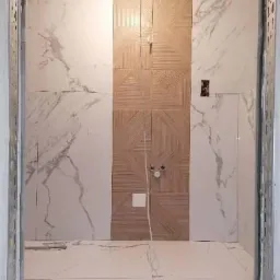 Ściana łazienki z częściowo ułożonymi płytkami: imitacja marmuru po bokach i drewnopodobny wzór geometryczny na środku, widoczne przyłącza wodne i puszka elektryczna, stan surowy wykończenia.