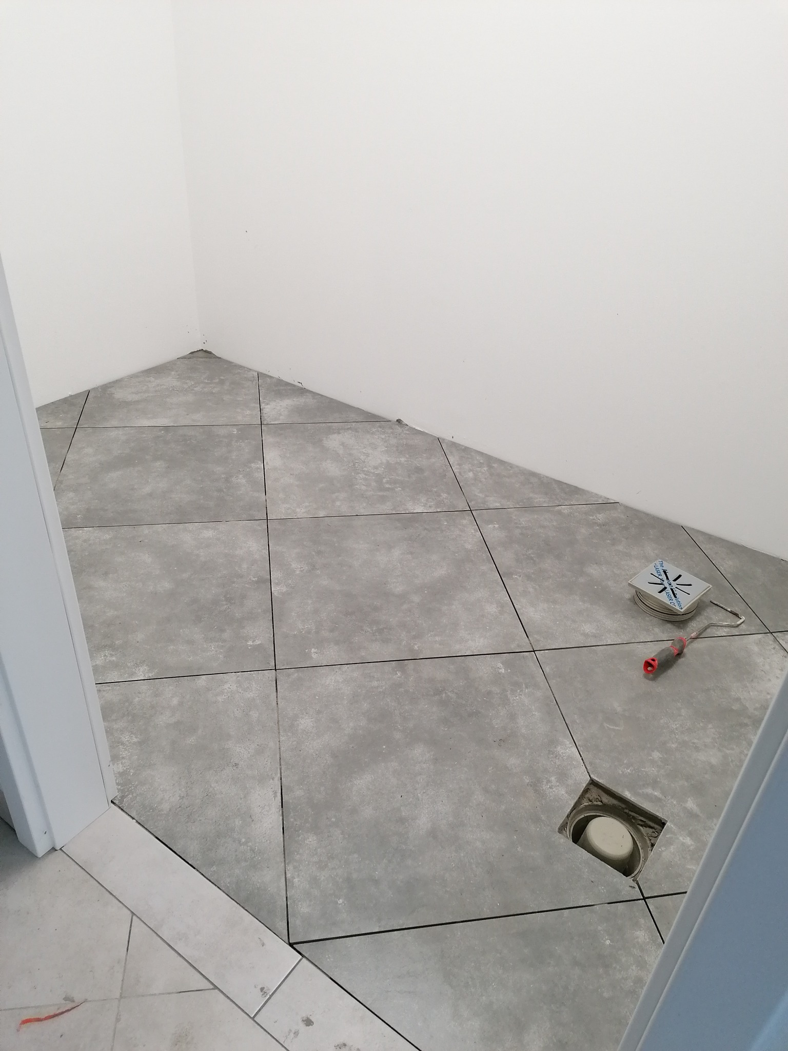 Szare płytki podłogowe w łazience ułożone pod skosem, widoczny odpływ liniowy i narzędzia glazurnicze