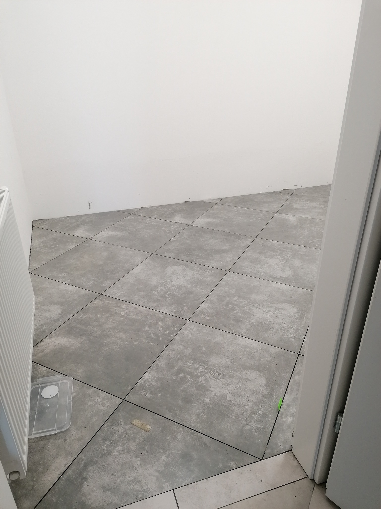 Szare, kwadratowe płytki podłogowe z fugami w świeżo wyremontowanym pomieszczeniu, widoczne białe ściany, grzejnik i fragment ościeżnicy drzwi, z plastikowym pojemnikiem na podłodze.