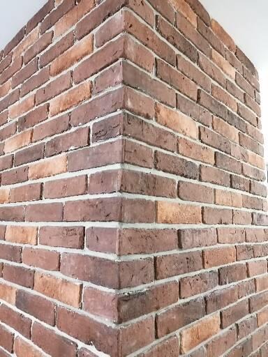 Fragment narożnej ściany wykończonej cegłą klinkierową w odcieniach czerwieni i brązu, z jasną fugą, widok z dołu.