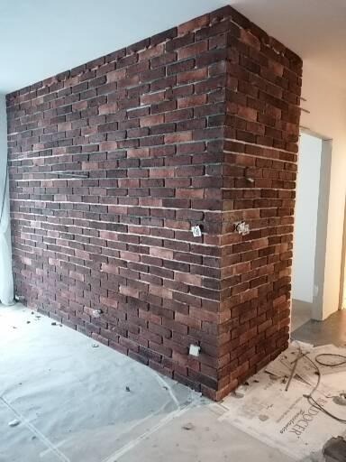 Ściana wyłożona dekoracyjną cegłą w odcieniach czerwieni i brązu, z widocznymi fugami i fragmentami instalacji elektrycznej, zabezpieczona folią ochronną na podłodze.