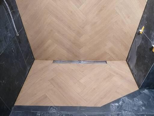 Ukończone ułożenie jasnych paneli podłogowych w jodełkę w łazience z czarnymi, marmurkowymi płytkami ściennymi i odpływem liniowym w podłodze.