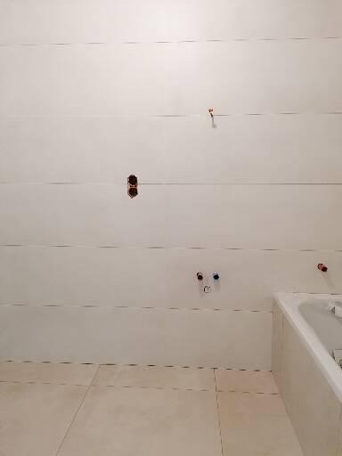 Surowa łazienka z ułożonymi płytkami podłogowymi i częściowo wykończoną ścianą z widocznymi przyłączami wodnymi i elektrycznymi przed montażem armatury.