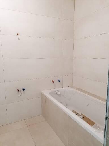 Wnętrze łazienki w trakcie wykańczania, z wanną obudowaną płytkami, widoczne przyłącza wodne na ścianie i świeżo położone jasne płytki na ścianach i podłodze.