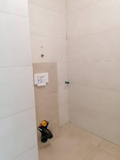 Wnętrze łazienki w trakcie wykańczania, widoczne białe płytki na ścianach i podłodze, częściowo wykończona ściana z instalacją wodną i workiem ze śmieciami, kartka z notatkami przyklejona do ściany.