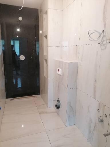 Nowoczesna łazienka w trakcie wykańczania, z białymi marmurkowymi płytkami, podtynkową instalacją WC i czarną kabiną prysznicową.