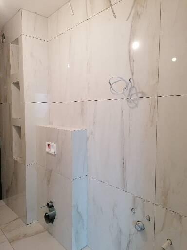 Łazienka w trakcie prac wykończeniowych: białe płytki imitujące marmur na ścianach, widoczne instalacje, częściowo zabudowana toaleta, podświetlone półki w ścianie.