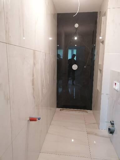 Wykończona łazienka z białymi, marmurkowymi płytkami na ścianach i podłodze, widoczne przyłącza wodne i ciemna szklana kabina prysznicowa.