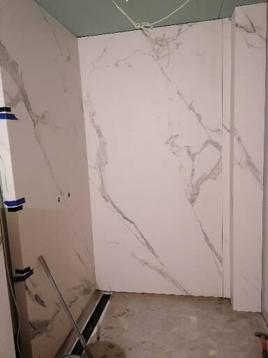 Ściany łazienki wyłożone płytami imitującymi marmur, widoczny odpływ liniowy w podłodze i fragment sufitu z płyt gipsowo-kartonowych w trakcie wykańczania.