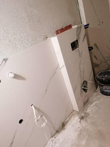 Płyty imitujące marmur na ścianie łazienki w trakcie montażu, widoczne otwory na instalacje, poziomica na górnej krawędzi, wiadro z masą szpachlową w tle.