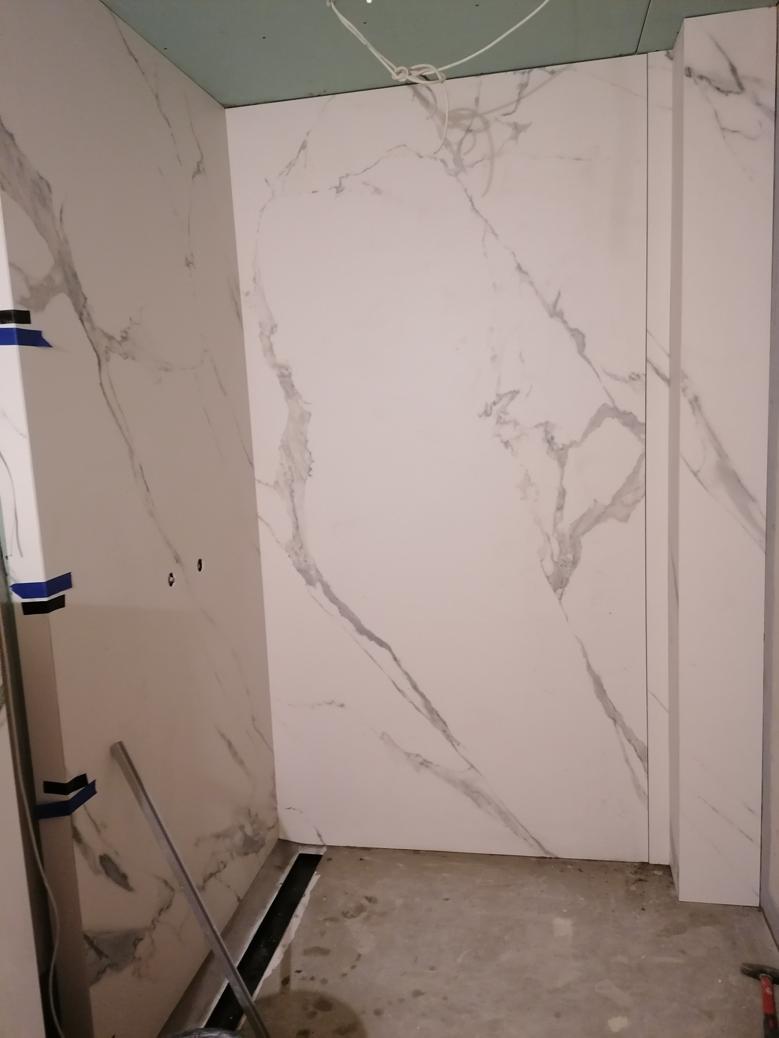 Ściany łazienki w trakcie wykańczania, wyłożone białymi płytami imitującymi marmur, widoczne narzędzia i elementy instalacji odpływowej w podłodze.