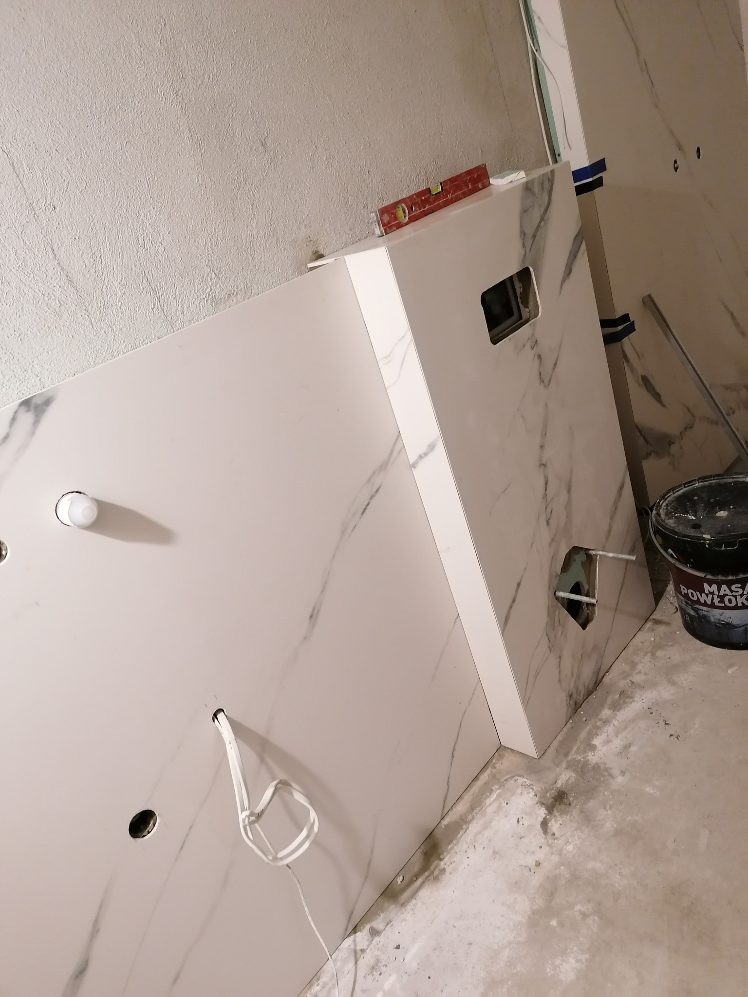 Płyty imitujące marmur z wyciętymi otworami na instalacje, ustawione w rogu pomieszczenia w trakcie prac wykończeniowych, widoczny poziomnica i wiadro z masą szpachlową.