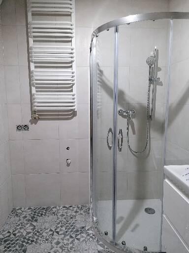 Nowoczesna kabina prysznicowa z chromowanymi detalami, biały grzejnik drabinkowy na ścianie z jasnych płytek, podłoga wyłożona wzorzystą mozaiką w odcieniach szarości.