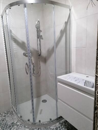 Kabina prysznicowa z chromowanymi profilami i biała umywalka z szafką w trakcie montażu, widoczne niedokończone wykończenie ściany z prawej strony.