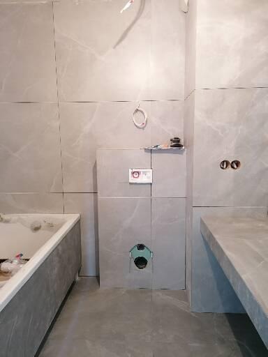 Szara łazienka w trakcie wykańczania: widoczne płytki imitujące marmur na ścianach, zabudowa podtynkowego WC, niedokończona zabudowa wanny, surowy blat umywalkowy, wystające kable elektryczne...