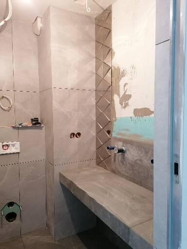 Szara łazienka w trakcie wykańczania, widoczne płytki imitujące marmur na ścianach, częściowo ułożona dekoracyjna mozaika w kształcie rombów, przygotowana instalacja wodna i elektryczna, surowy...