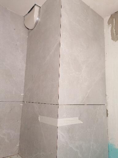 Szare płytki imitujące marmur na ścianie w trakcie montażu, z widoczną taśmą maskującą i fugami, fragment narożnika z obudową rury.