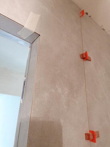 Szare płytki ścienne w trakcie montażu, z użyciem krzyżyków dystansowych i taśmy maskującej przy ościeżnicy.