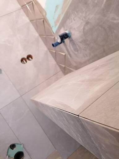 Ściana łazienki wyłożona szarymi płytkami imitującymi marmur, z widocznymi otworami na gniazdka i wystającą rurą, fragment blatu pod umywalkę z ułożonych płytek, w tle ślady zaprawy.