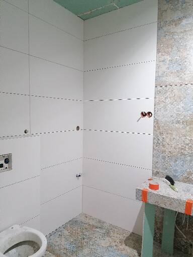 Przygotowanie ścian w łazience do położenia płytek, widoczne płyty gipsowo-kartonowe z zaznaczonymi liniami, częściowo położone płytki dekoracyjne, narzędzia na blacie i fragment toalety.