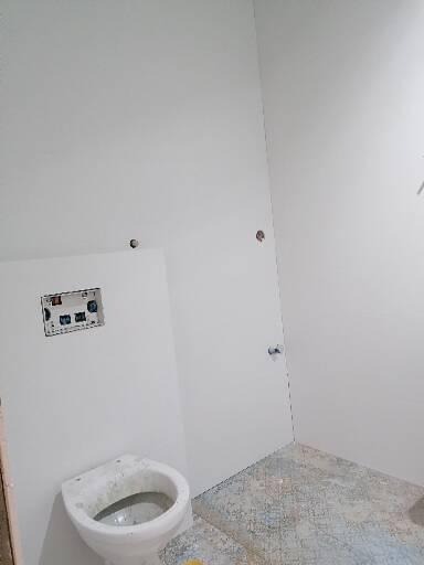 Surowa łazienka w trakcie prac: białe ściany z widocznymi otworami na instalacje, częściowo położone płytki podłogowe w odcieniach szarości i błękitu, oraz zamontowana, ale jeszcze nieoczyszczona...