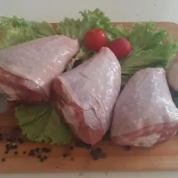 Trzy surowe udka indycze z widoczną skórą i mięsem, ułożone na drewnianej desce z liśćmi sałaty, dwoma pomidorkami cherry, główką czosnku, gałązką pietruszki i ziarnami pieprzu.