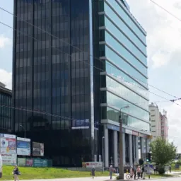 Nowoczesny biurowiec z ciemną szklaną fasadą i jasną szklaną fasadą odbijającą chmury, widoczny z poziomu ulicy z billboardami i przechodniami.