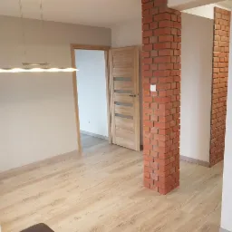 Jasne wnętrze po remoncie z drewnopodobną podłogą, ceglanymi filarami, nowoczesną lampą i dwiema drewnianymi ościeżnicami z drzwiami.