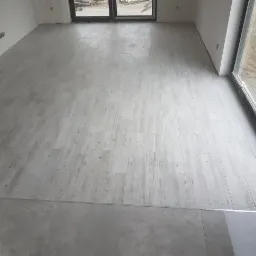 Jasnoszara podłoga wyłożona panelami imitującymi drewno, widoczne przejście do płytek ceramicznych w odcieniu szarości, duże okno z widokiem na zewnątrz w stanie surowym.