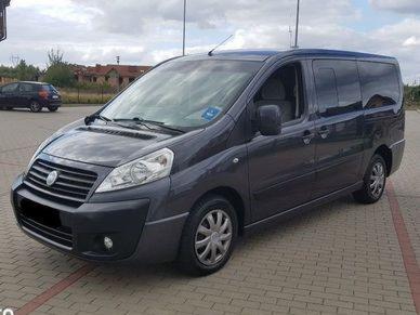 Ciemnoszary bus osobowy na kostce brukowej w Pabianicach, widok z boku, zarys budynków w tle.