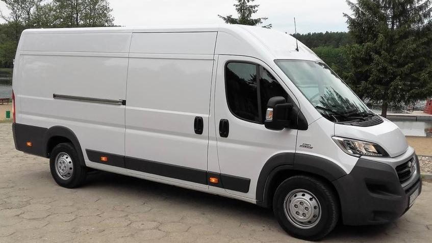 Biały bus dostawczy Fiat Ducato z wysokim dachem, zaparkowany na betonowym placu, widok z boku.