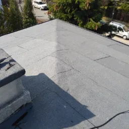 Widok z góry na płaski dach pokryty papą, z widocznymi łączeniami i niewielkimi pęknięciami, obok komina leżą narzędzia, w tle zabudowa jednorodzinna i drzewa.