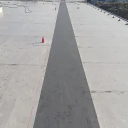 Szeroki, szary pas świeżo nałożonej izolacji na płaskim dachu przemysłowym z widocznymi świetlikami i elementami mocującymi.