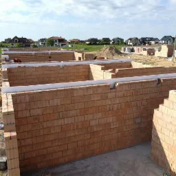 Wznoszone mury fundamentowe z czerwonej cegły ceramicznej z białym ociepleniem na obrzeżach, widok na plac budowy z domami w tle.