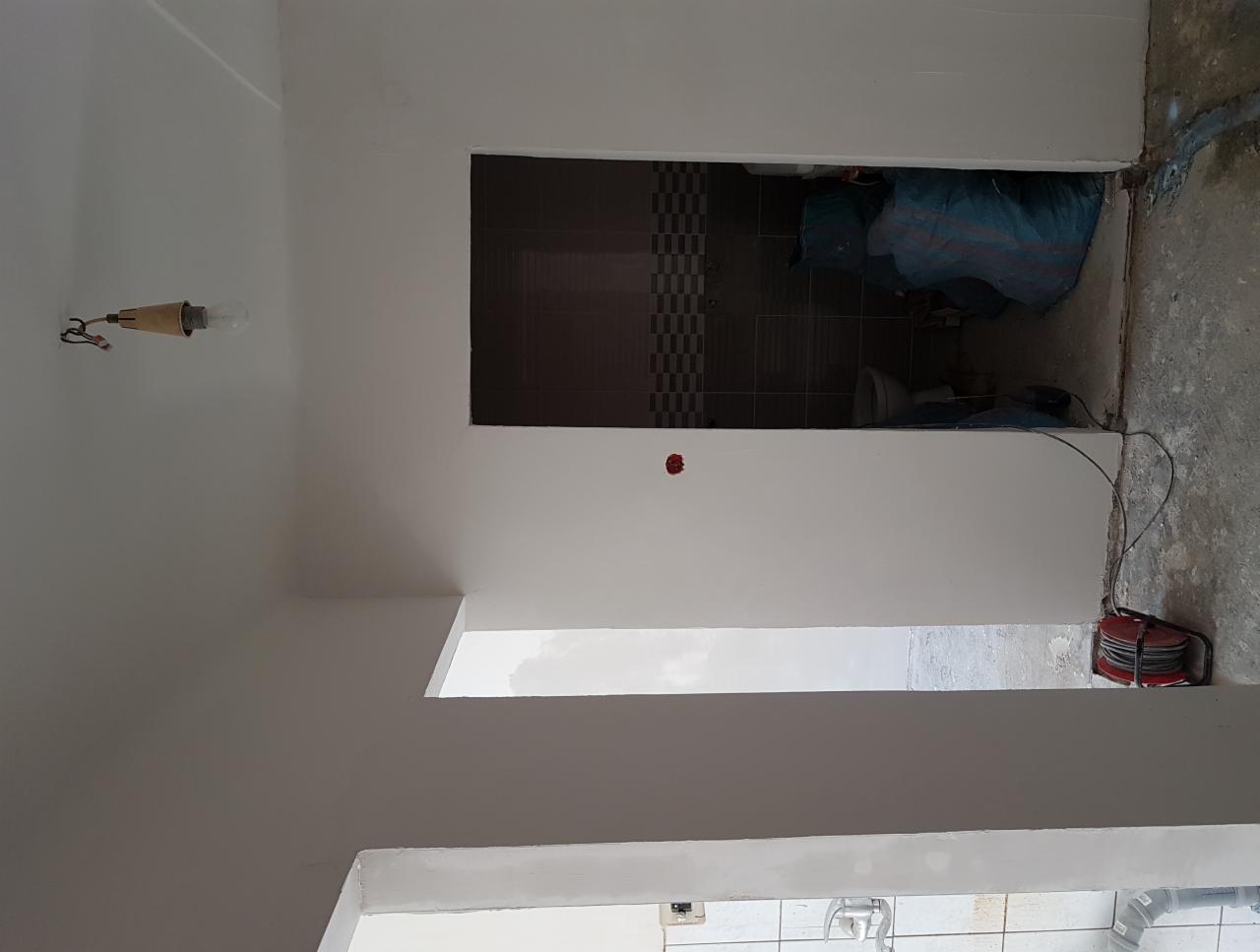 Wnętrze w trakcie remontu: widoczne świeżo pomalowane ściany, surowy beton na podłodze, wystająca żarówka bez klosza i fragment łazienki z szarymi płytkami w tle, z rozłożonym kablem elektrycznym...