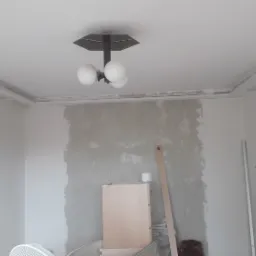 Pokój w trakcie prac wykończeniowych: ściany częściowo pokryte gładzią, listwy sufitowe przygotowane do malowania, widoczna lampa sufitowa i elementy wyposażenia wnętrza.