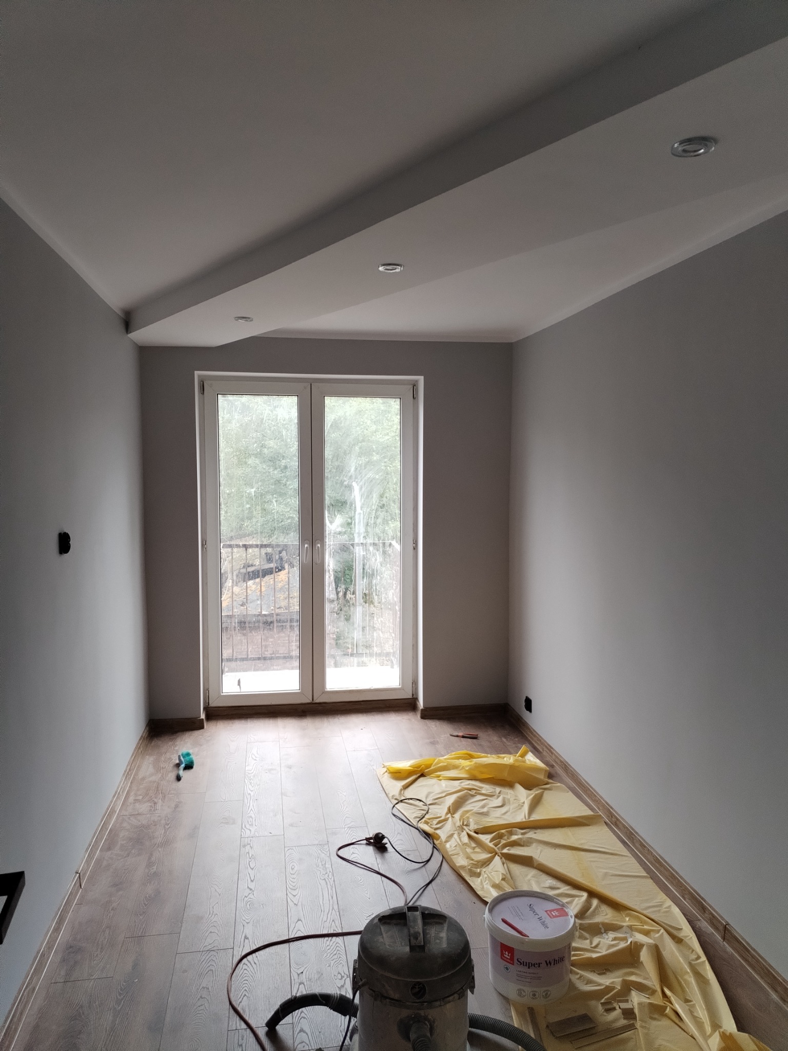 Pokój po malowaniu ścian na szaro, z widocznym wiadrem farby Super White, folią ochronną na podłodze i szlifierką do gładzi na parkiecie.