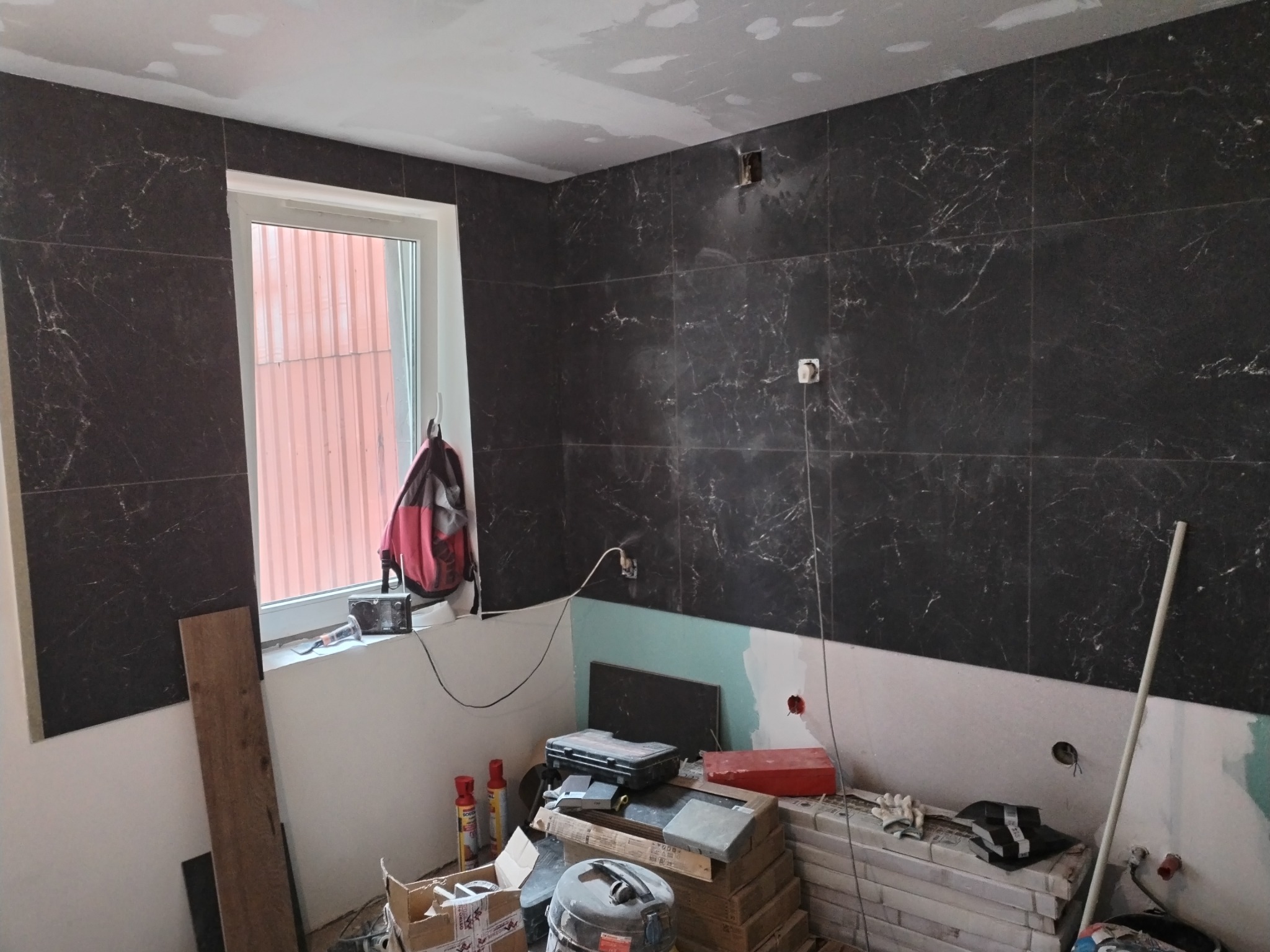 Układanie ciemnych płytek imitujących marmur w łazience, widoczne narzędzia i materiały budowlane w trakcie pracy.